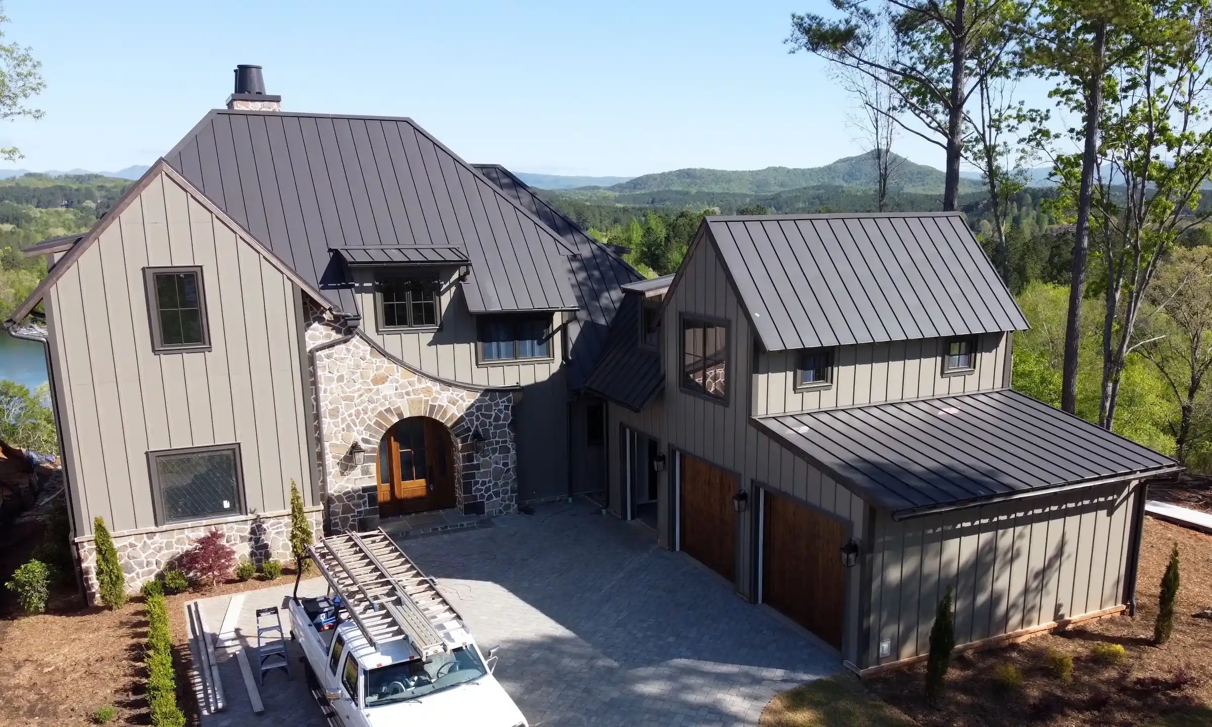 Front view of a custom-built home with vertical metal siding and a dark standing seam metal roof, featuring a stone archway entry and a multi-car garage, surrounded by mountains and forest.